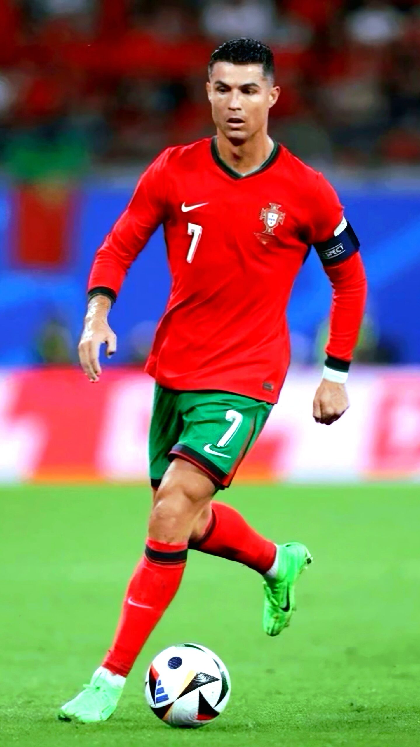 A male soccer player in a red and green Portugal uniform with the number 7 controls a ball on the field, wearing green shoes and a black captain's armband. The blurred background shows fans and flags, ideal for streaming on Romania IPTV.