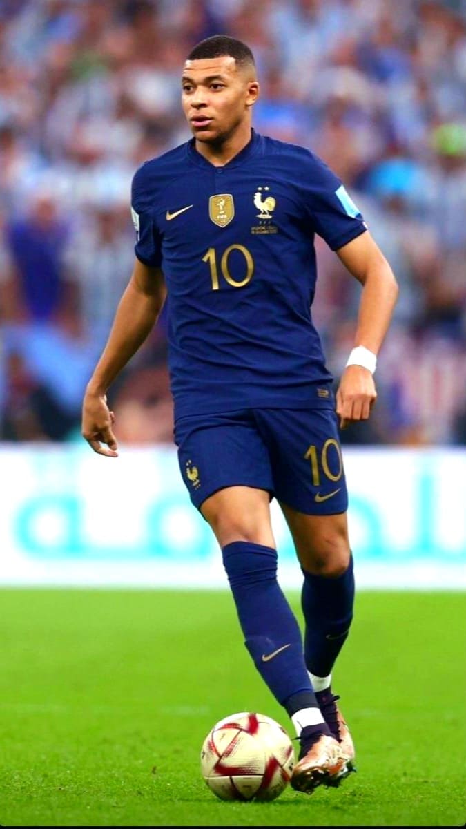 A male soccer player in a dark blue France national team jersey with number 10 dribbles a ball on a green field during a match, as viewers enjoy the action via Romania IPTV, with a blurred crowd in the background.