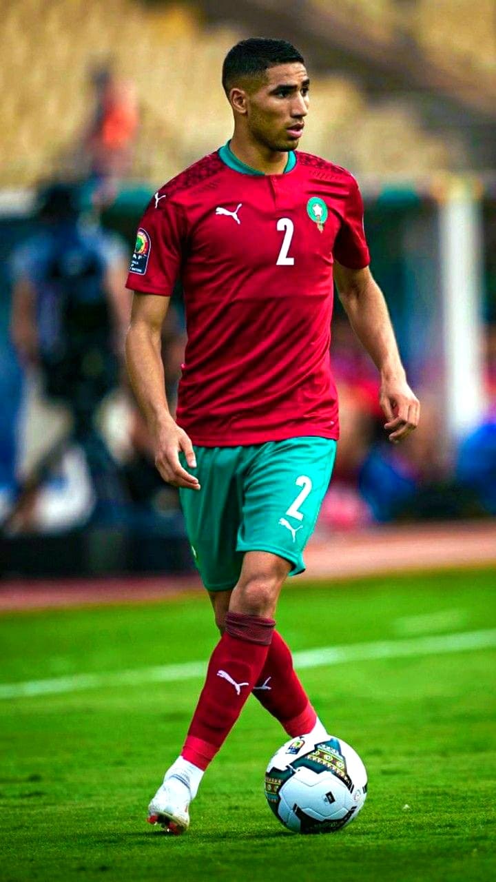 A male soccer player in a red and green Morocco national team uniform, wearing number 2, controls a soccer ball during a match. The action, visible to Romania IPTV viewers, unfolds on the field with a blurred stadium and spectators in the background.