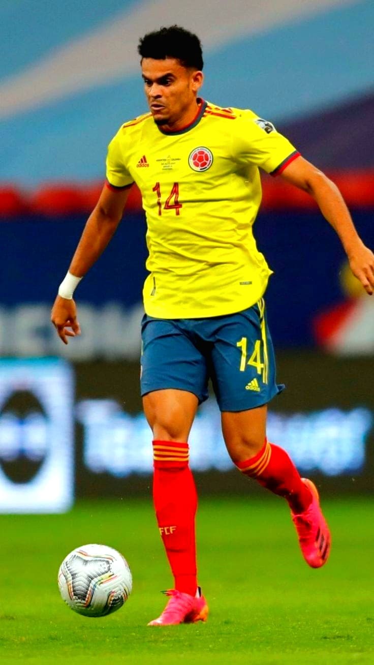 A male soccer player in a yellow Colombia national team jersey with number 14 dribbles a soccer ball on the field during a match, wearing blue shorts, red socks, and pink cleats—action you can stream live via Romania IPTV.
